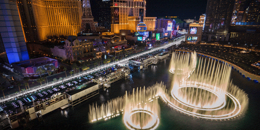 Aerial night view of the Las Vegas Strip, featuring the illuminated Bellagio Fountain dancing in front of bright hotels and casinos, with colorful lights reflecting on the water.