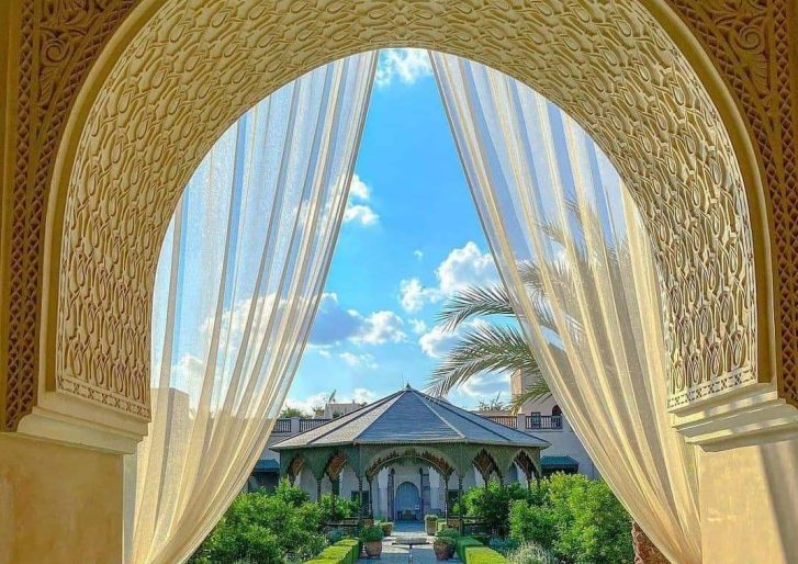 View through an ornate arched doorway with sheer curtains revealing a lush garden, palm trees, and a pavilion under a bright blue sky with scattered clouds. Intricate patterns decorate the arch.