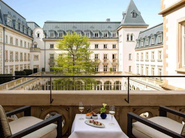 A picturesque view of a courtyard from a balcony, featuring elegant historical architecture. In the foreground, a table is set with plates of appetizers, a glass of white wine, and a plant, surrounded by two cushioned chairs.