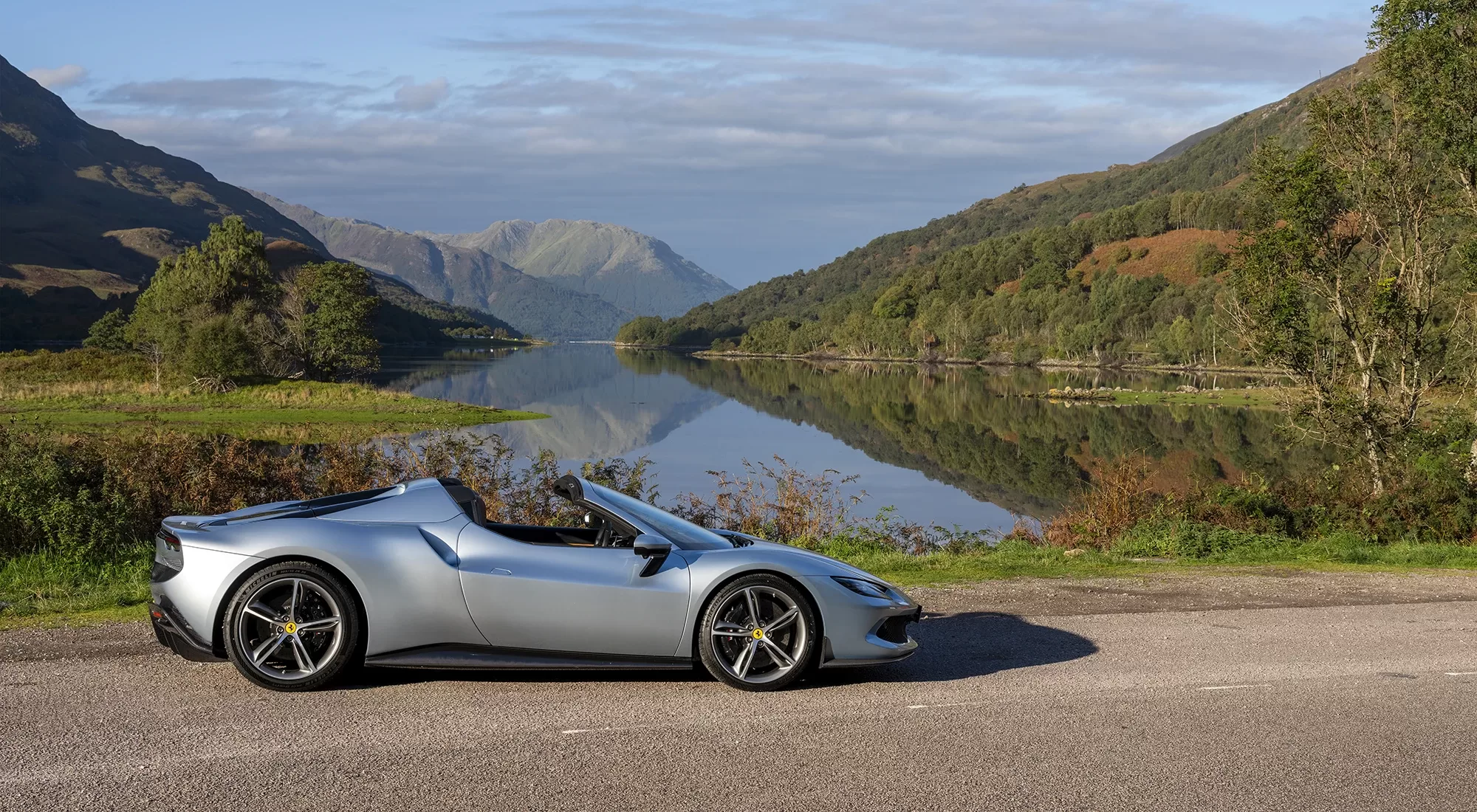 A sleek silver convertible sports car parked beside a calm lake, with mountains and green hills reflected in the water under a partly cloudy sky.