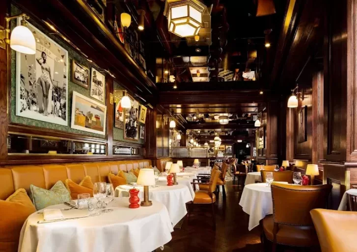 A cozy, elegant restaurant with brown leather chairs, round tables with white tablecloths, warm lighting, and framed photos on dark wood-paneled walls. The atmosphere is inviting and upscale.
