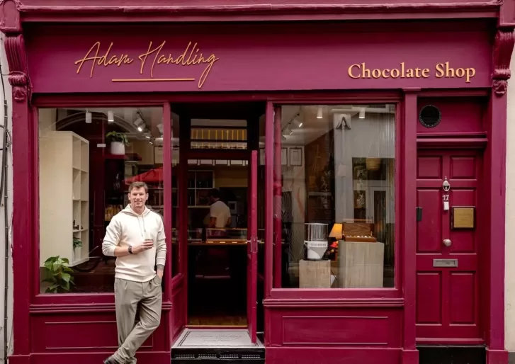 A man in a light sweater and khaki pants stands, holding a drink, outside a maroon storefront that reads Adam Handling Chocolate Shop with large windows displaying chocolates inside.