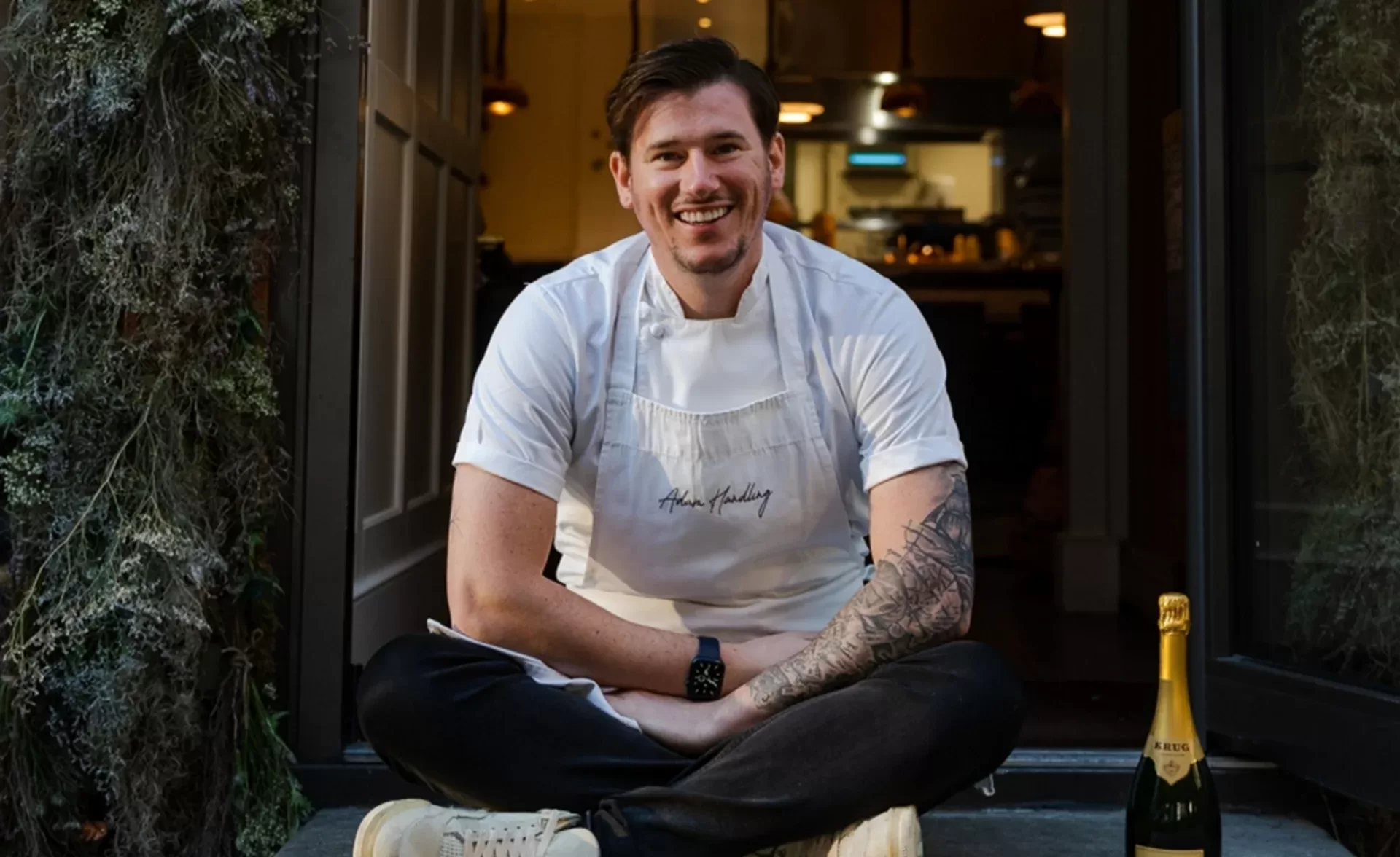 A smiling chef in a white apron sits cross-legged in a doorway, with a kitchen visible behind him. He has short hair, a tattooed arm, and a bottle of sparkling wine placed on the ground nearby.