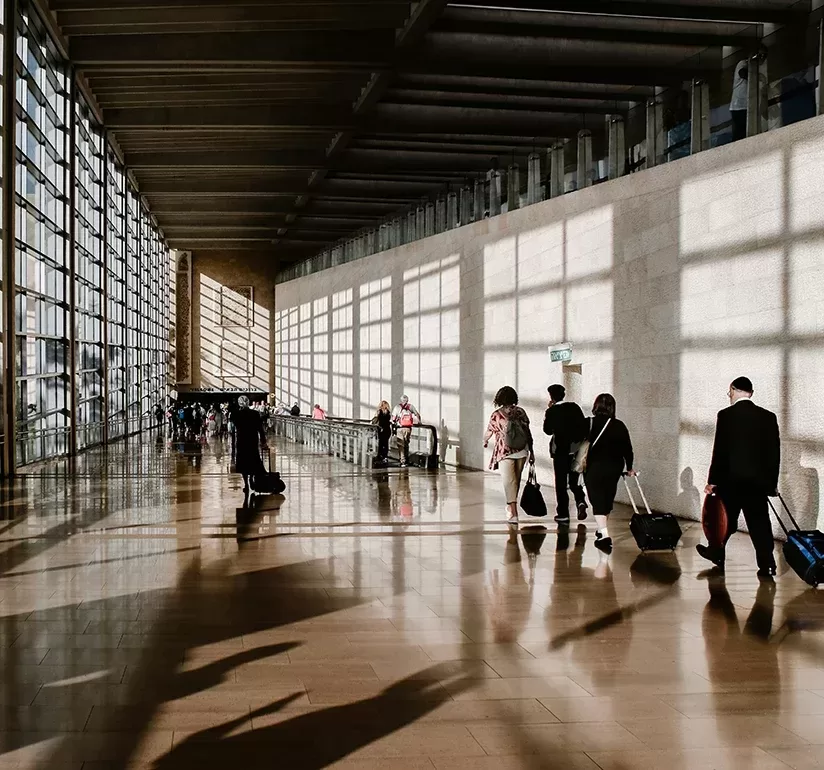 People walk with suitcases through a sunlit airport terminal, casting long shadows on the shiny floor. The space is lined with large windows on one side and a textured wall on the other.