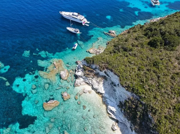 Aerial view of a rocky coastline with lush green vegetation on the right. Crystal-clear turquoise waters display visible rocks beneath the surface. Several boats, including a large yacht, are anchored near the shore.