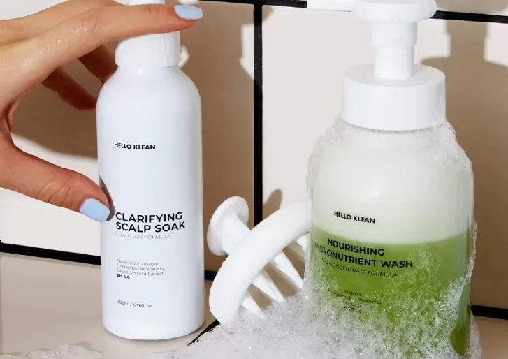 A hand holding a white bottle labeled Clarifying Scalp Soak next to a pump dispenser with green liquid labeled Nourishing Micro-Nutrient Wash. Both products are on a tiled surface with bubbles around them.