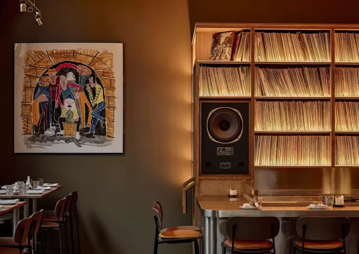 A cozy restaurant interior with a large painting on the wall, shelves filled with vinyl records, a prominent speaker, and neatly set tables and chairs under warm lighting.