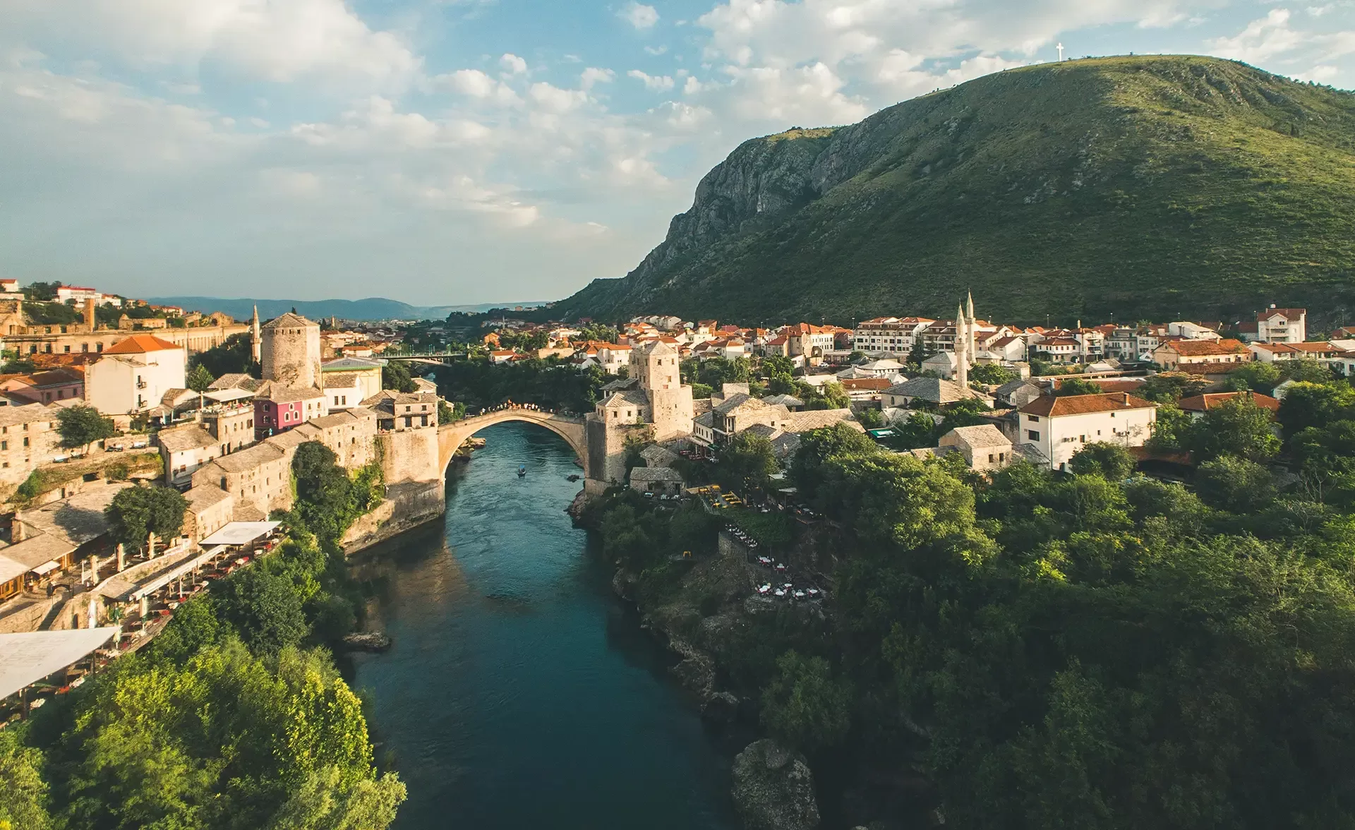 Aerial view of a historic stone bridge arching over a calm river, surrounded by a town with red-roofed buildings. A lush landscape and a large hill provide a scenic backdrop under a partly cloudy sky.