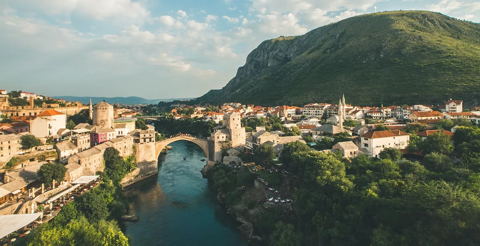 Aerial view of the Stari Most, a historic stone bridge, arching over a river in Mostar, Bosnia and Herzegovina. Surrounded by lush greenery and rooftops under a partly cloudy sky, with a large hill in the background.