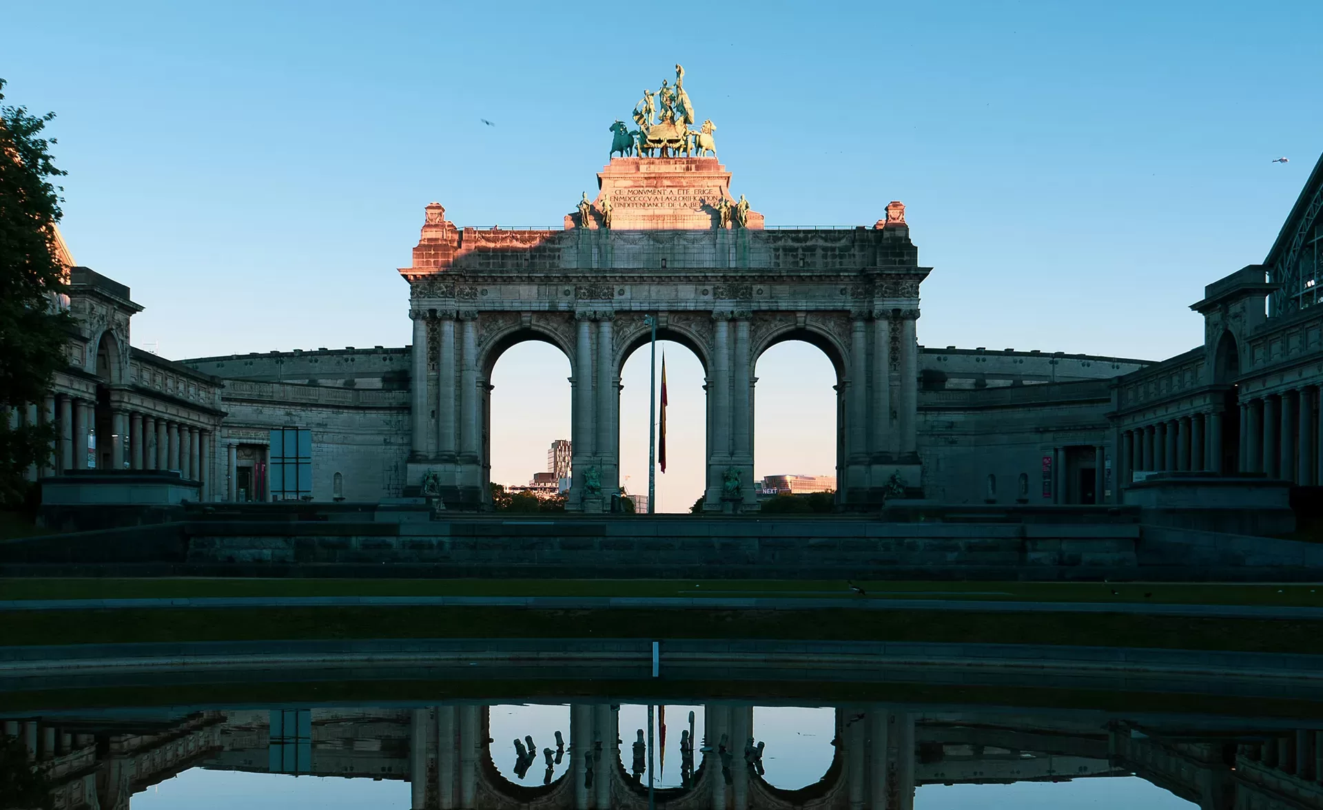 A majestic arched monument with a chariot sculpture atop is silhouetted against the sky at sunset. The structure is mirrored in a calm pool below, surrounded by symmetric buildings and trees under a clear blue sky.