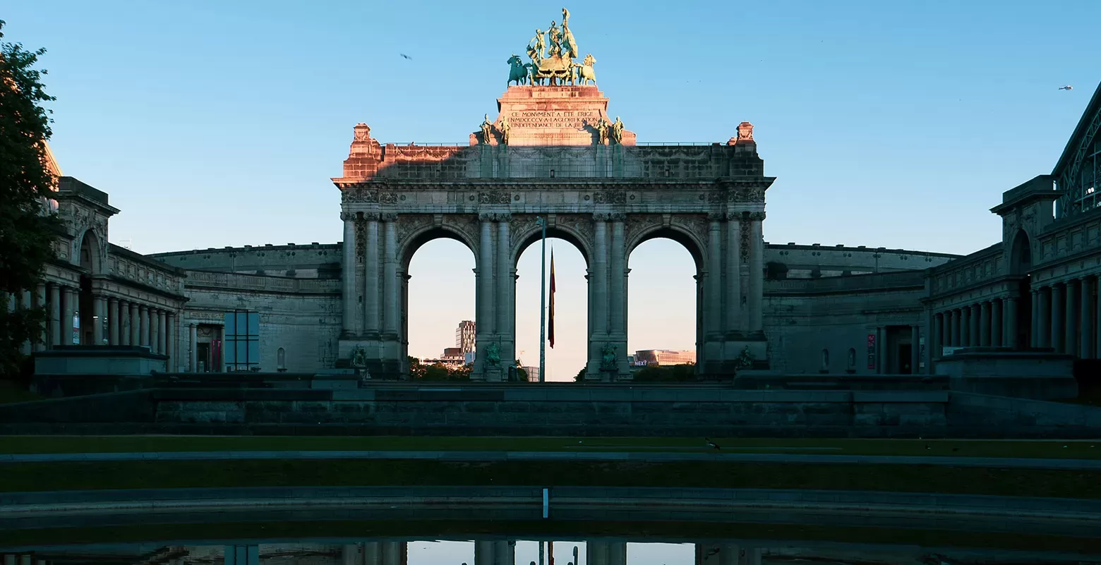 The image shows the Cinquantenaire Arch in Brussels, Belgium, at sunset. The arch features three arches and a bronze quadriga statue on top. The sky is clear, and the foreground includes a reflective pool of water.
