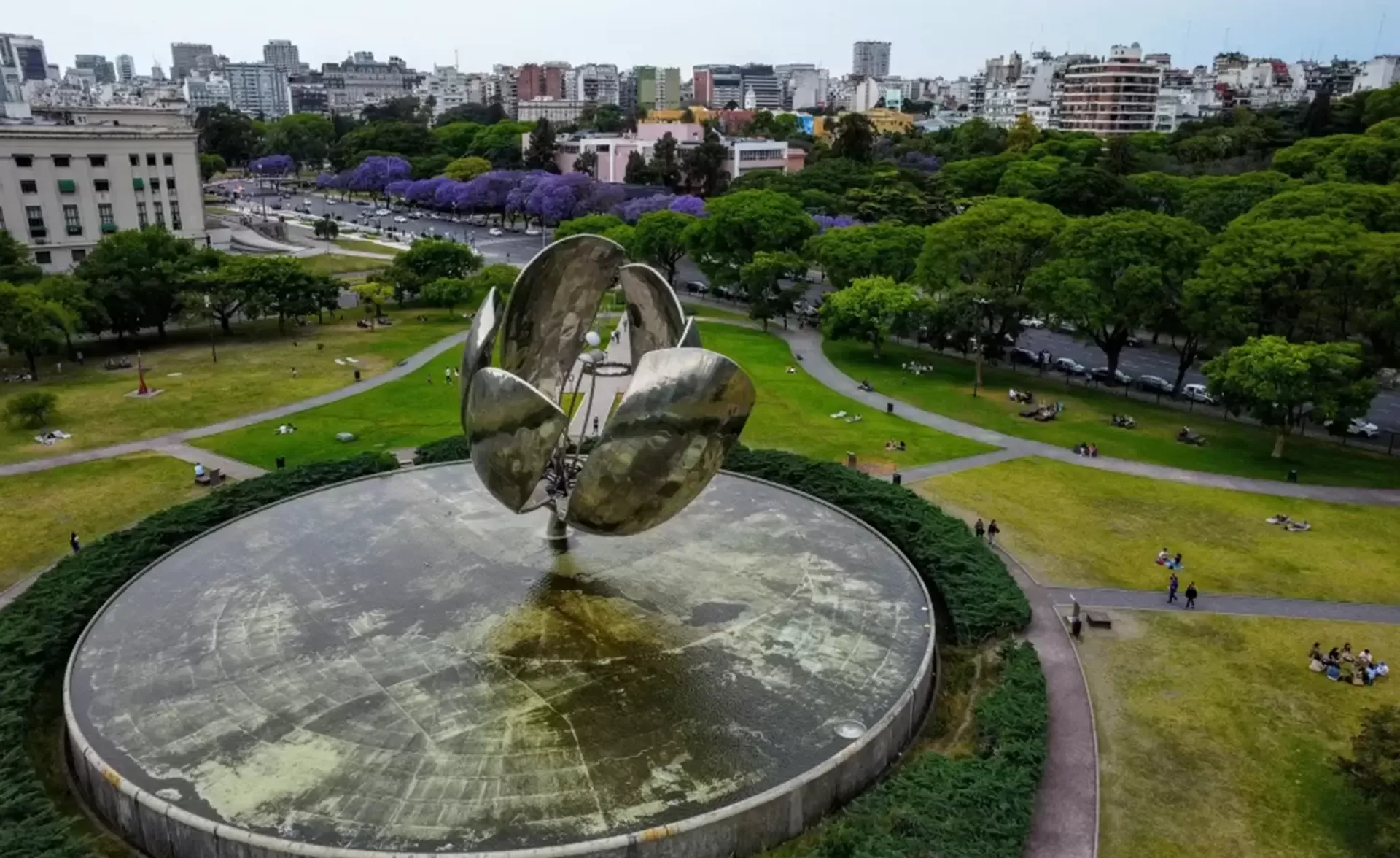 Aerial view of a large metallic flower sculpture in a park, surrounded by green lawns and trees. People are sitting on the grass, and a city skyline is visible in the background.