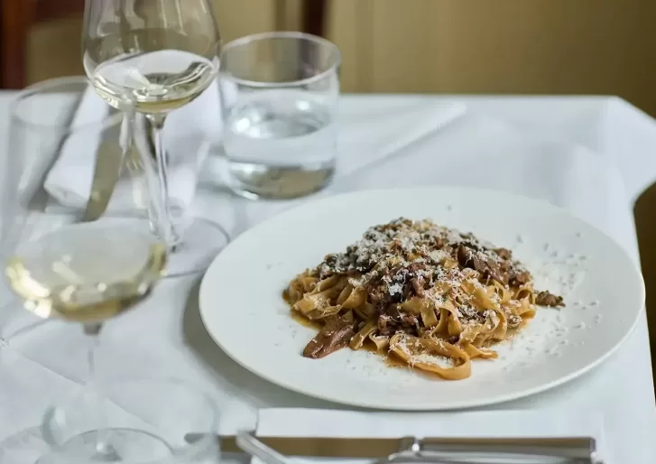 A plate of tagliatelle pasta with meat sauce and grated cheese sits on a white tablecloth, accompanied by two glasses of white wine and a glass of water, with cutlery and a napkin nearby.