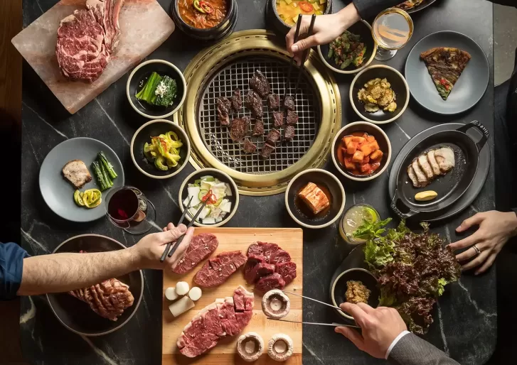 Top view of a Korean BBQ meal, featuring a grill with meat cooking, surrounded by side dishes, raw meats, and hands holding chopsticks and cutting meat. There are plates of vegetables, sauces, and drinks on a dark table.