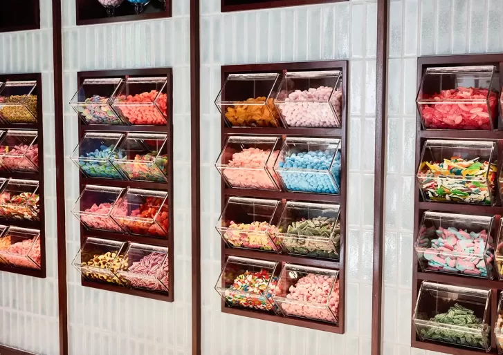 Clear plastic bins filled with various colorful candies, including gummies and licorice, are neatly arranged on a tiled wall in a self-serve candy shop.