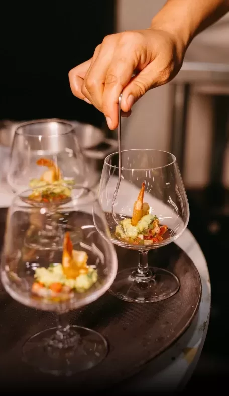 A hand carefully places a garnish in a glass containing a colorful appetizer, likely a shrimp cocktail with avocado and diced vegetables. The dish is elegantly presented in three stemmed glasses on a dark tray.