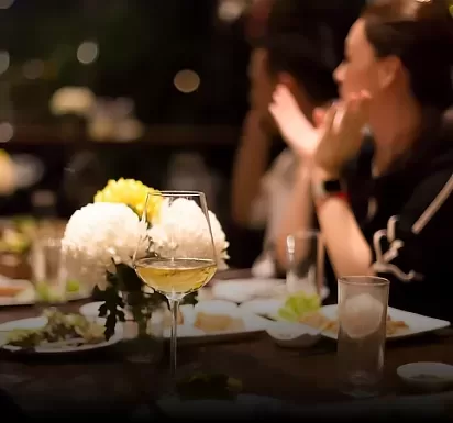 A softly lit dining table with a glass of white wine, white flowers, and plates. In the background, blurred figures are engaged in conversation, creating a warm and inviting atmosphere.