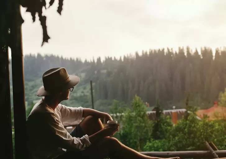A person wearing a hat and glasses sits outdoors with a cup in hand, gazing at a forested landscape during sunset or sunrise. Lush green trees and distant hills are visible under a soft, golden sky.