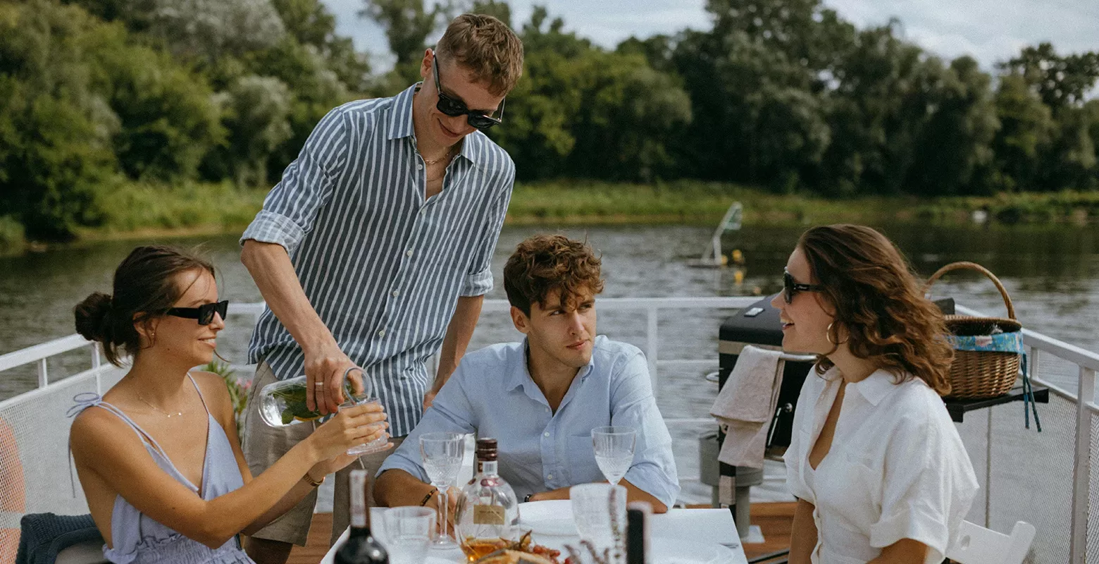 Four people sit around a table on a boat, enjoying drinks together. One person is pouring a beverage for another. They are all wearing sunglasses and light summer clothes, with a river and greenery in the background.