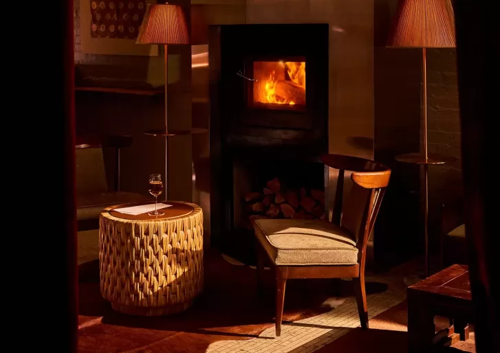 A cosy room with a lit fireplace, a wooden chair, a round woven table with a glass of wine, and two standing lamps casting a warm glow. Logs are stacked below the fireplace.
