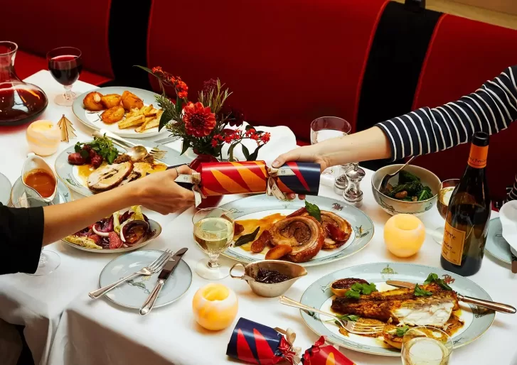 A festive dinner table with two people pulling a Christmas cracker. Plates feature roasted meat, vegetables, and a side dish. The table is decorated with candles, flowers, and a glass of white wine at Ham Yard Hotel, Soho in London.