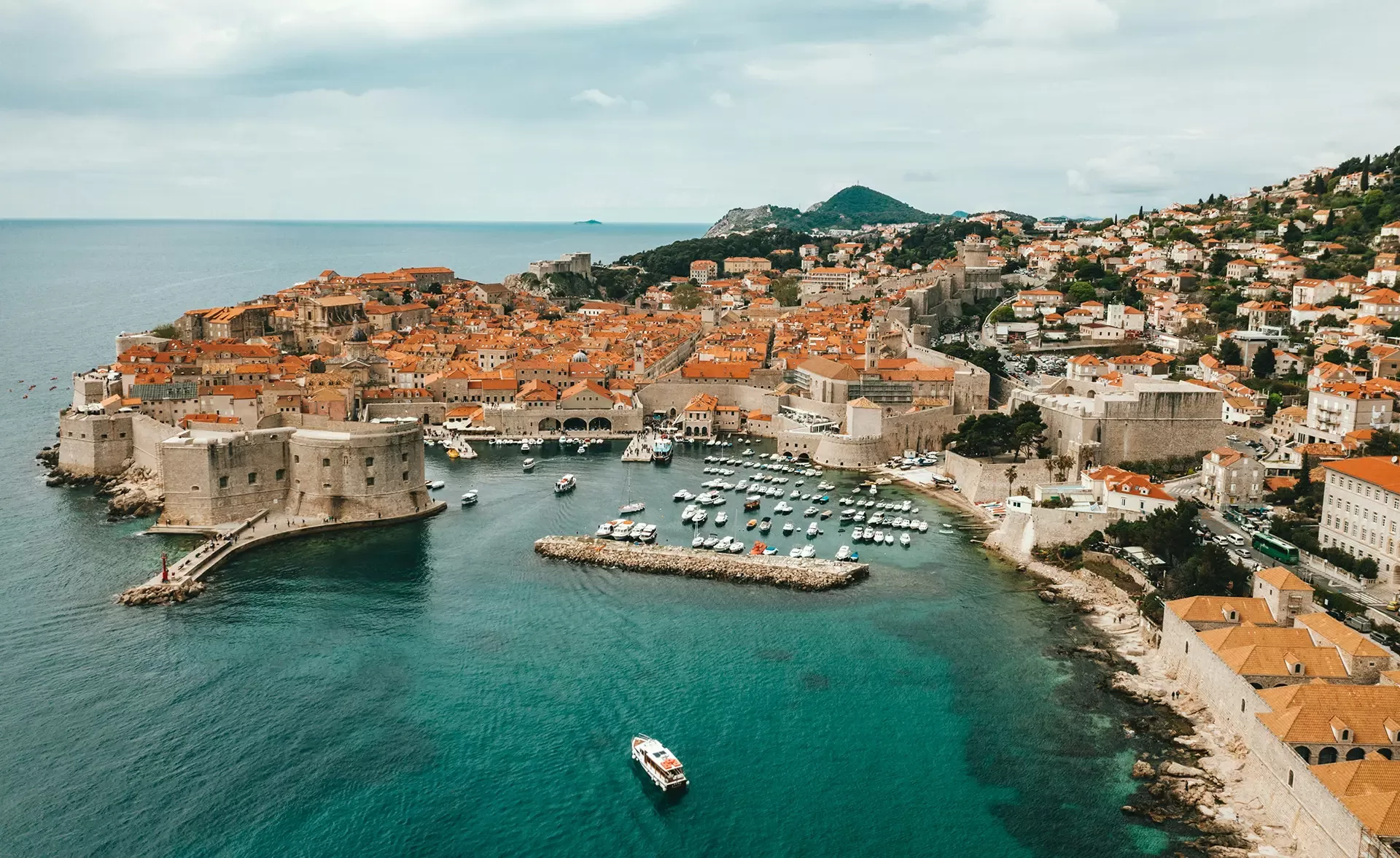 Aerial view of a coastal city with historic, orange-roofed buildings surrounded by medieval walls. A marina with boats is nestled in the turquoise waters. A small boat navigates the open sea, with lush hills in the background.