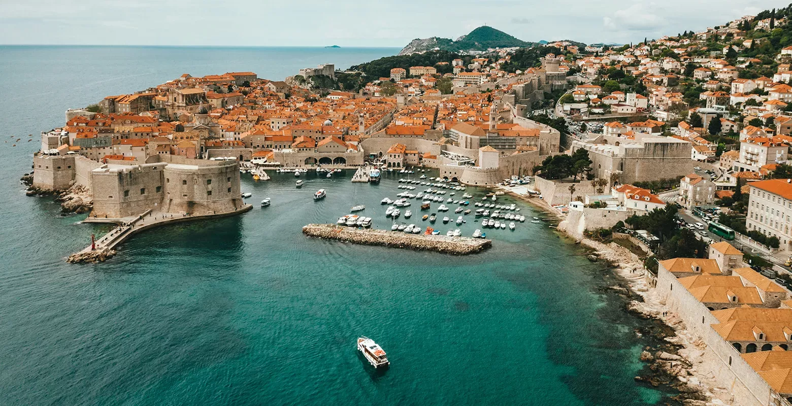 Aerial view of a coastal city with a historic fortress and orange-roofed buildings by the sea. Numerous boats are docked in the harbor, and a single boat is moving in the turquoise water. Hills and greenery surround the city.