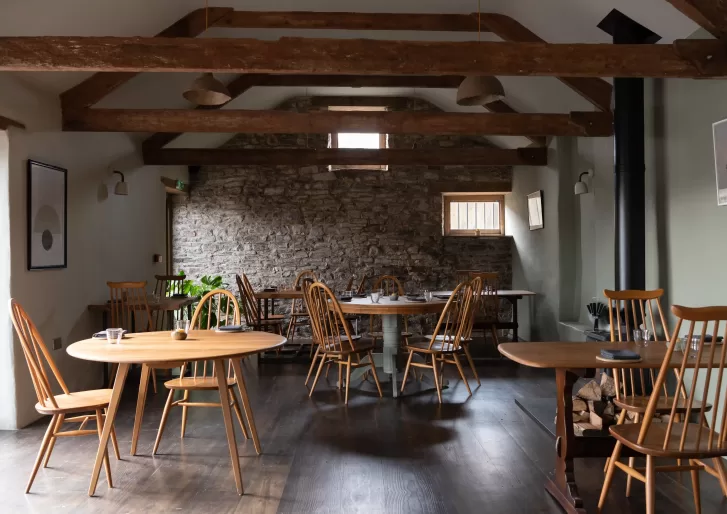 Quint cottage style restaurant interior with Cornish stone walls and round wooden tables under wooden beamed roof at Crocadon Farm, Cornwall