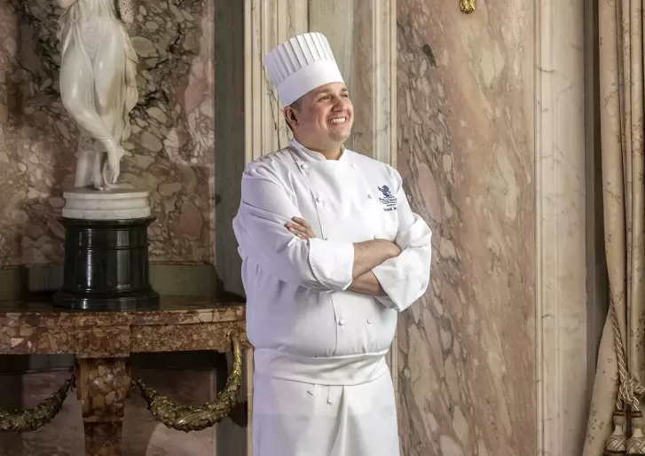 A chef in a white uniform and tall hat stands with arms crossed, smiling, in an elegant room with marble walls and a classical statue in the background.