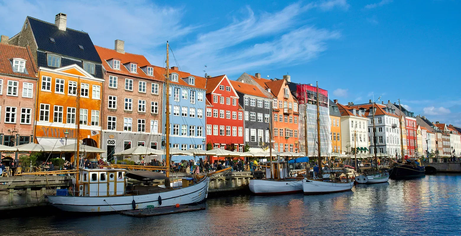 Colorful historic buildings and moored boats line the Nyhavn waterfront canal in Copenhagen, Denmark, under a clear blue sky.