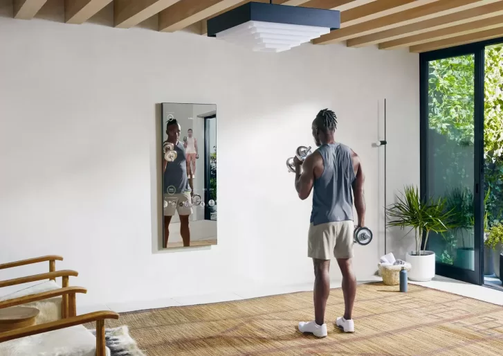 Man in gym clothes with weights working out opposite a home fitness technology mirror in a sunny room