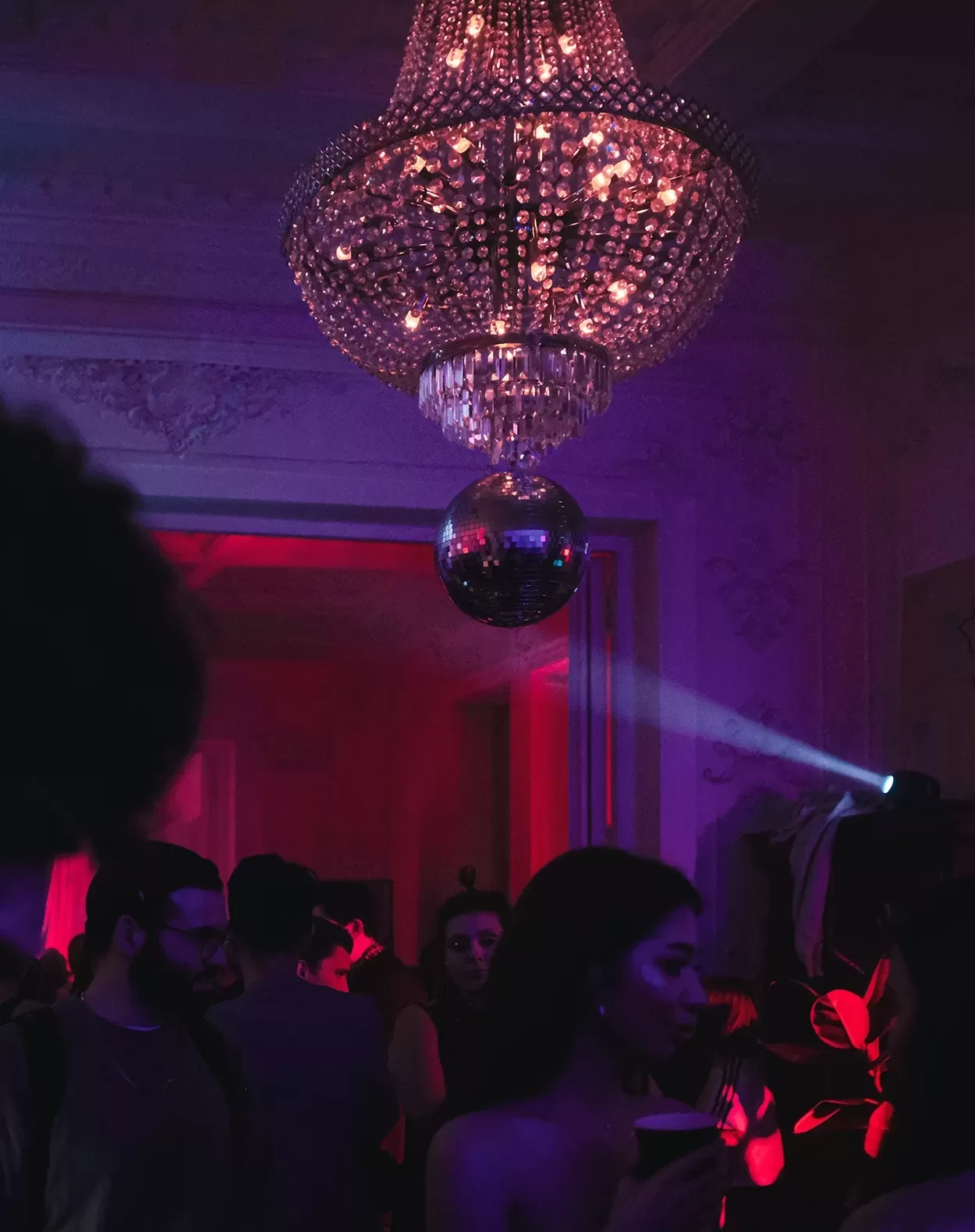 A dimly lit party scene with people mingling under a large, ornate chandelier and a mirrored disco ball. Purple and pink lighting creates a vibrant atmosphere. A narrow beam of light shines onto the disco ball, casting reflections around the room.