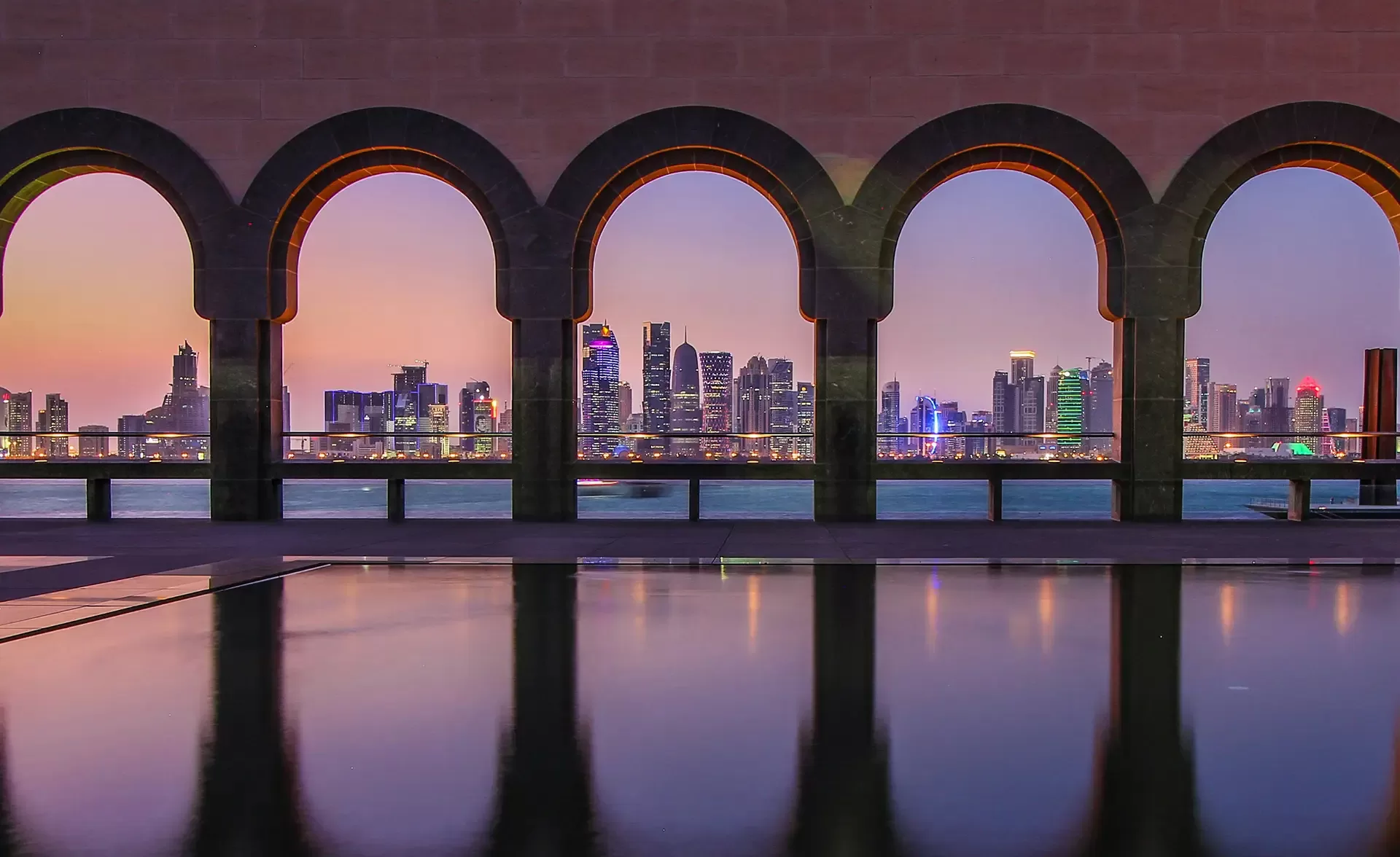 View of a city skyline at sunset through five arched windows. The calm water in the foreground reflects the arches, while the buildings in the background are illuminated with colorful lights.