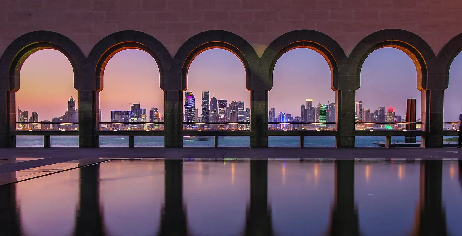 A series of arched windows frames a vibrant, illuminated city skyline at dusk. The foreground features a reflective pool, enhancing the colorful, serene ambiance of the scene.