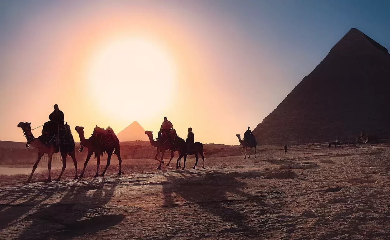 A group of people ride camels silhouetted against a sunset, with the pyramids of Giza in the background. The scene creates long shadows across the desert landscape, highlighting the grandeur of the pyramids under a clear sky.