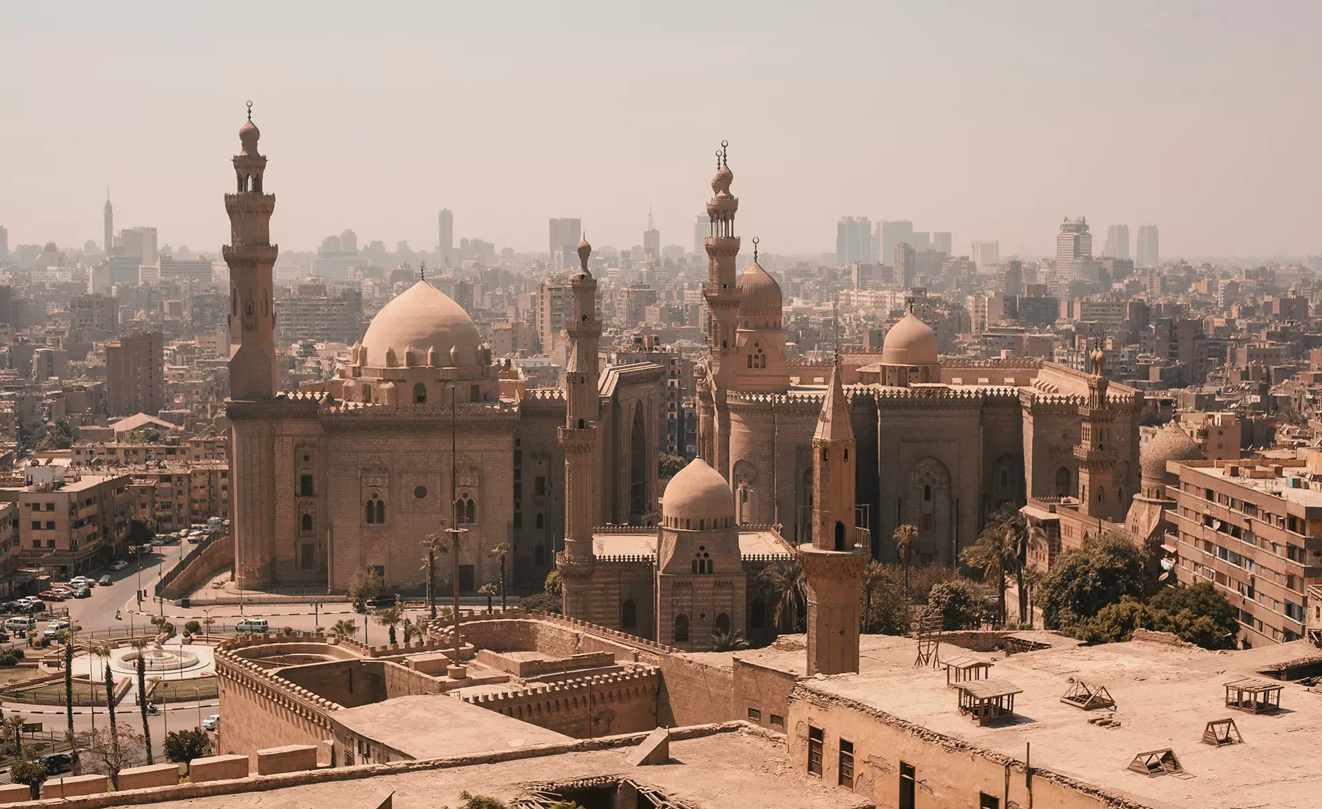 A panoramic view of Cairo with historic Islamic architecture. Prominent domes and minarets rise amidst tan brick buildings, set against a backdrop of a sprawling modern cityscape under a hazy sky.