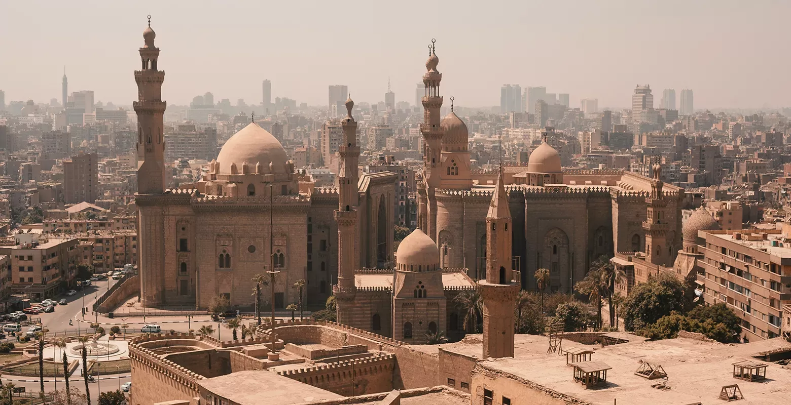 A panoramic view of Cairo showcasing the historic Sultan Hassan Mosque and the Al-Rifai Mosque. The citys dense urban landscape and modern high-rises are visible in the background under a hazy sky.
