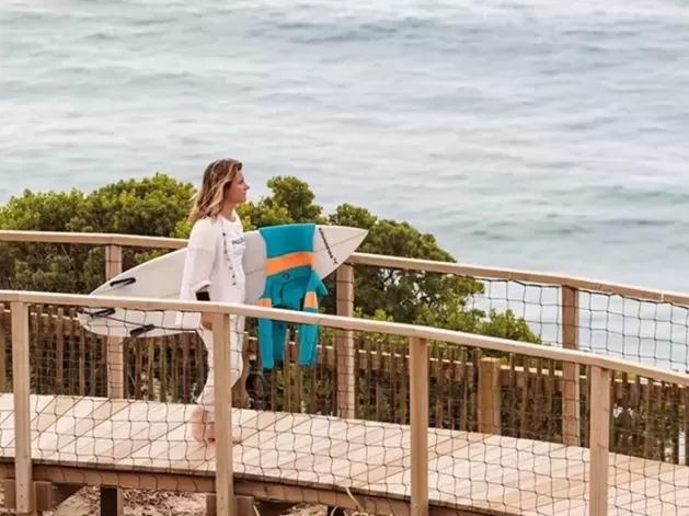Women stood on a wooden walkway bridge across beach next to the sea holding a surf board at Ericeira, Portugal