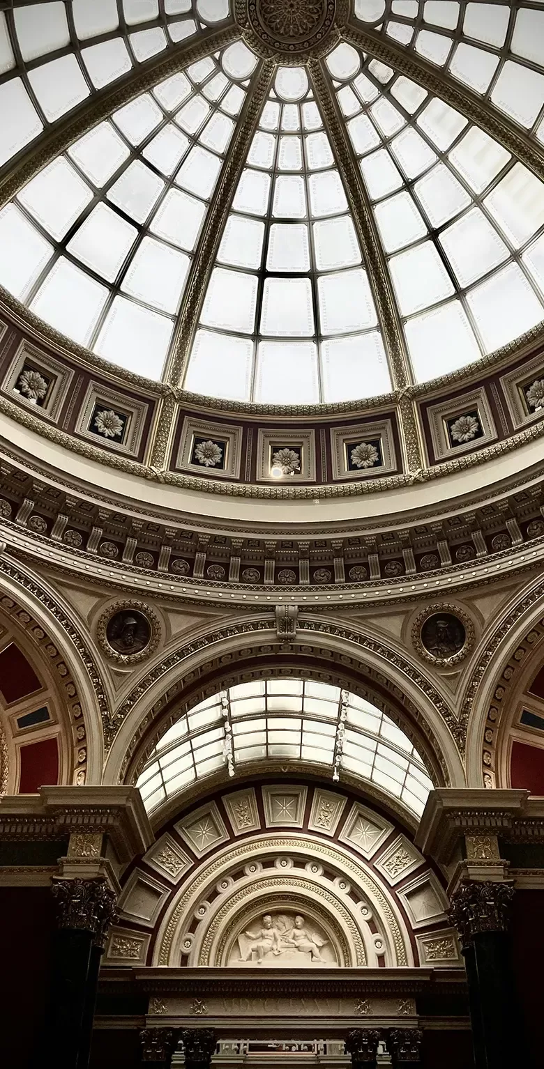 A grand, ornate domed ceiling with a large, segmented glass skylight and detailed architectural carvings in a historic building, featuring arches, columns, and intricate decorative elements.
