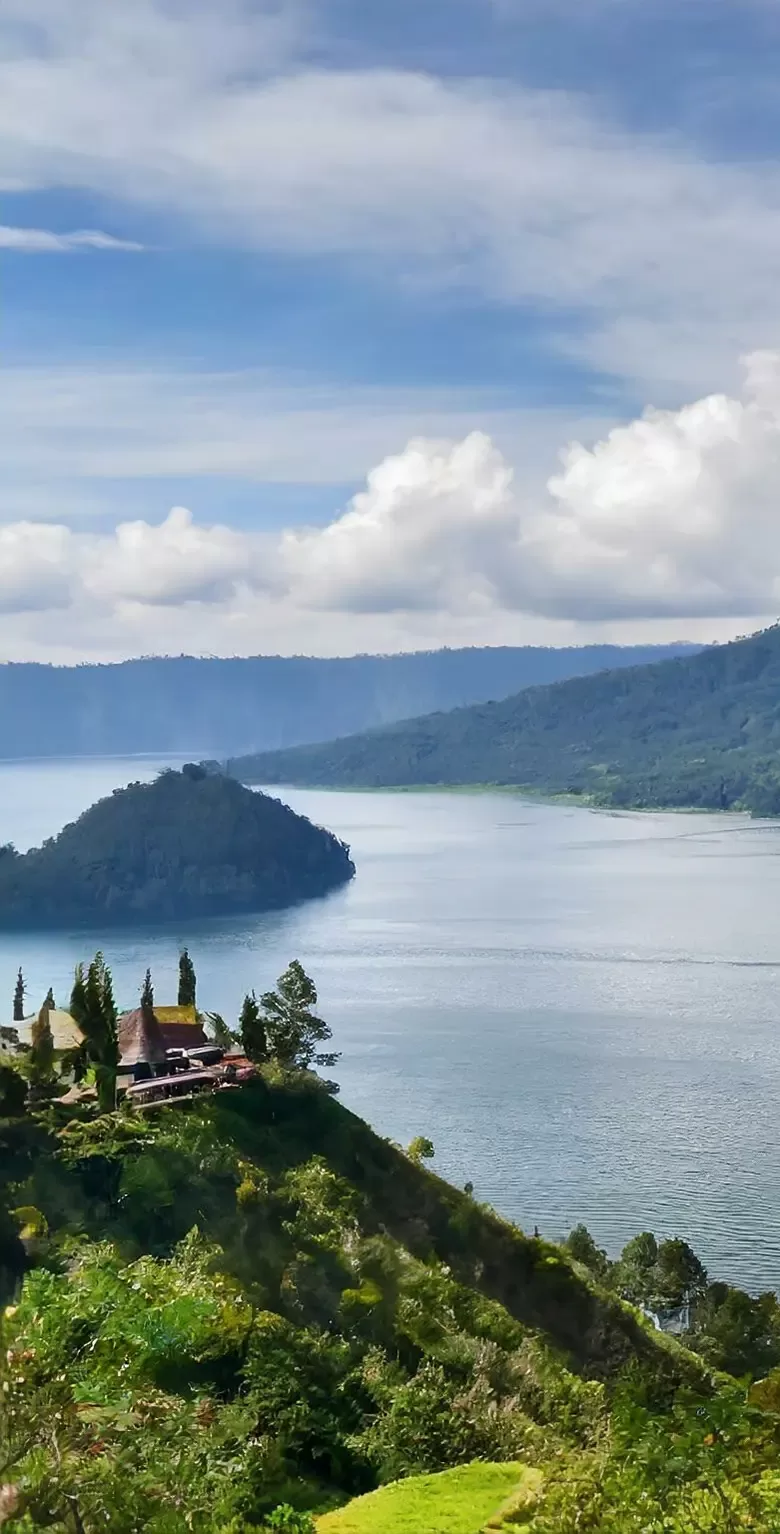 A serene lake surrounded by lush green hills under a partly cloudy blue sky, with a cluster of buildings and trees on the foreground hillside.
