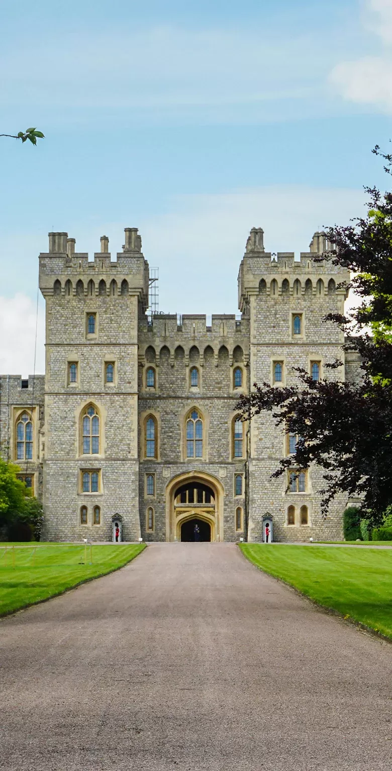 A wide stone castle with tall towers, arched windows, and a large entrance stands at the end of a long, straight path bordered by green lawns and trees under a blue sky.