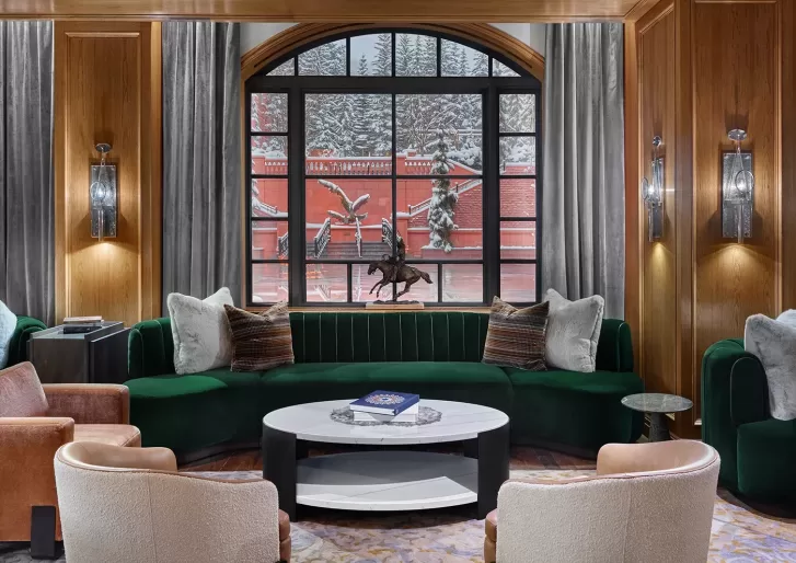 A cozy living room with a green velvet sofa, two beige chairs, a round white coffee table, and large window showcasing a snowy outdoor scene with red brick walls and trees.