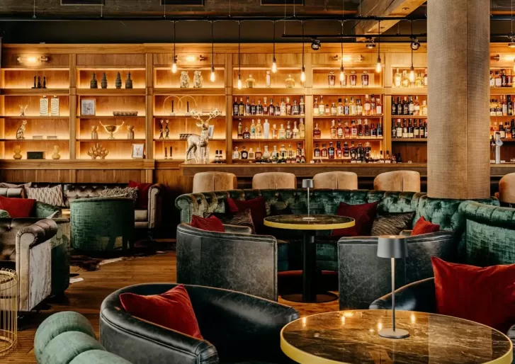 A cozy lounge with leather chairs and plush sofas surrounds round tables. A well-lit wooden bar is lined with a variety of bottles, creating a warm and inviting atmosphere. Soft lighting enhances the ambiance.