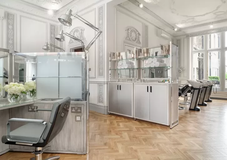 A modern, elegant hair salon features styling chairs, large mirrors, overhead lights, and glass shelves with towels and products. The space has ornate white walls, tall windows, and a herringbone wood floor.