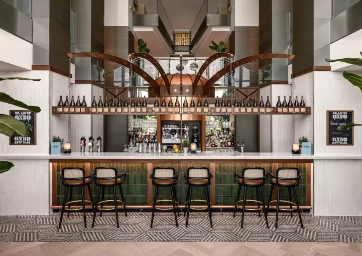 Modern bar interior with a long counter lined by six chic bar stools. Shelves stocked with bottles are displayed above, flanked by two chalkboards. The flooring has a geometric pattern, and large plants decorate the space.