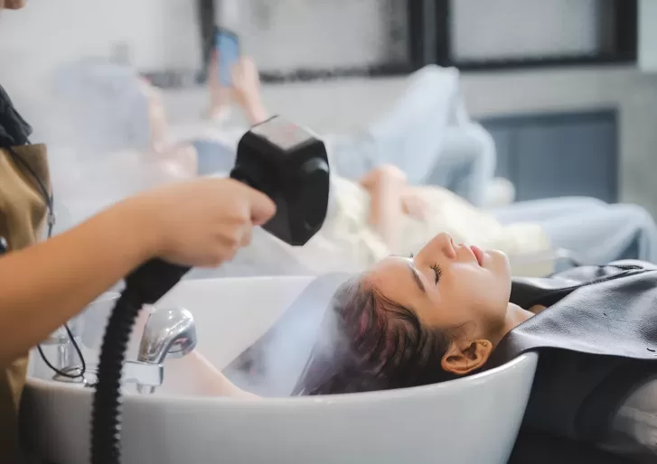 A person relaxes at a salon with their head in a sink while a stylist steams their hair using a handheld device. Another person is blurred in the background, sitting and using a phone.