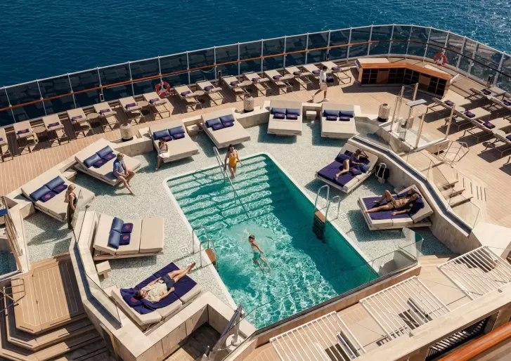 A view from above of a cruise ship’s pool deck, with people relaxing on lounge chairs and swimming in a small pool surrounded by clear railings and the ocean in the background.