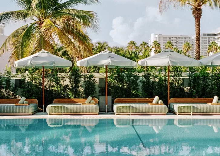 Poolside scene with lounge chairs and white umbrellas lined up along a tranquil pool. Palm trees and lush greenery are in the background, with buildings visible in the distance under a partly cloudy sky.