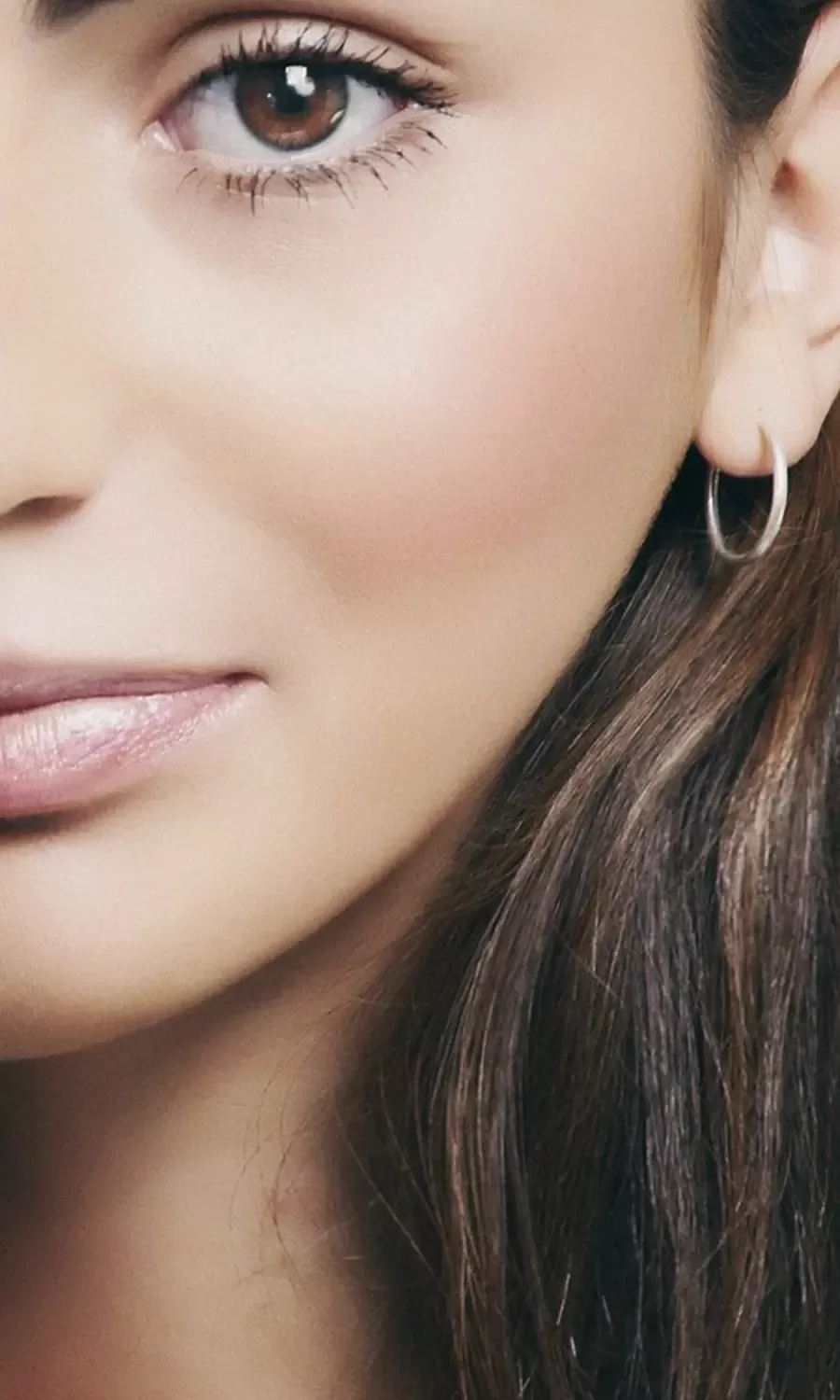 Close-up of a womans face and hair, showing her brown eye, smooth skin, natural makeup, light pink lips, and a small silver hoop earring.
