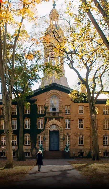 A person walks toward a historic brick building adorned with ivy. The building features a central clock tower. Tall trees with autumn foliage surround the pathway leading to the entrance.